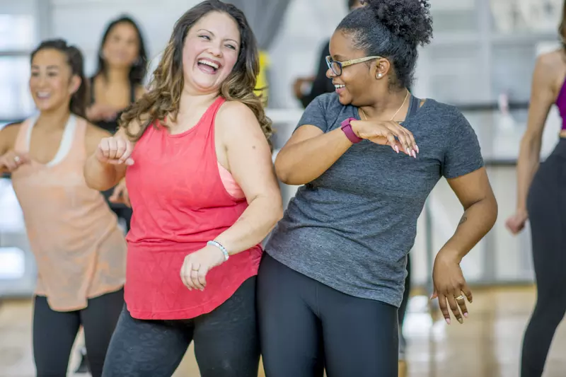 A group of adult women in an exercise class, with two women at the forefront of the shot bumping hips.