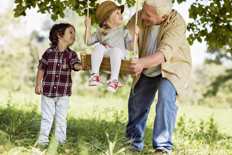 A grandfather with his two grandkids, one sitting on a wood swing, the other helping their grandfather push the swing.