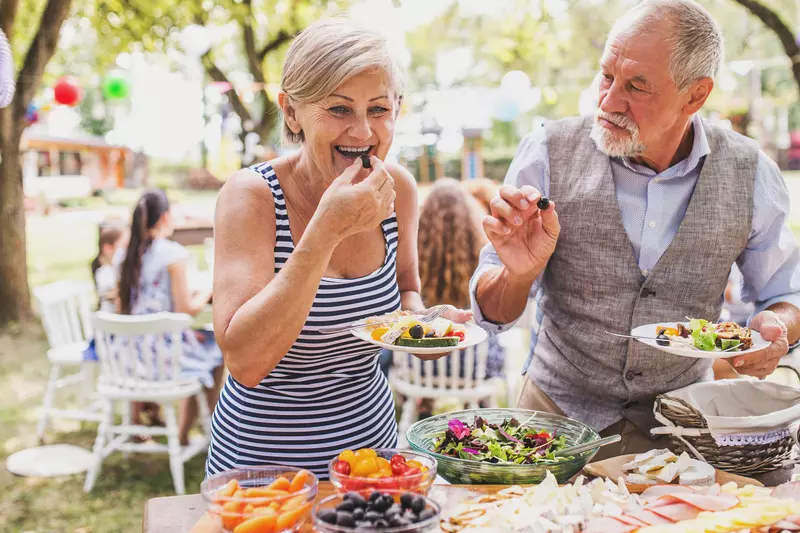 A senior couple at an outdoor party enjoys a heart healthy lunch together. They are standing eating over a buffet table.
