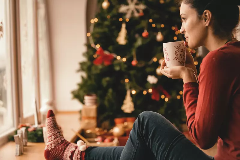 Woman sitting on the ground next to a Christmas tree, drinking something from a mug.