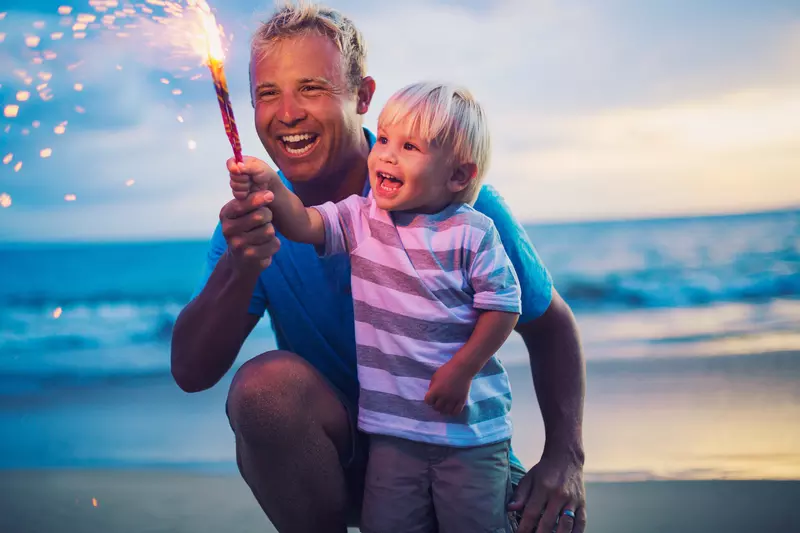 A father and son holding a firework on the beach at sunset.