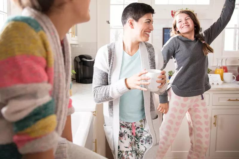 A woman dancing in the kitchen with her daughters and drinking coffee.
