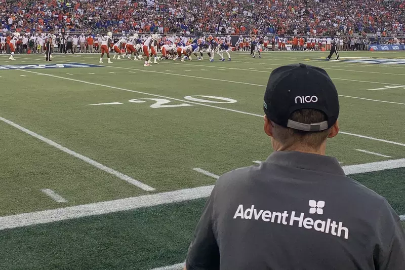 Man wearing shirt with AdventHealth logo on the sidelines of a football game.