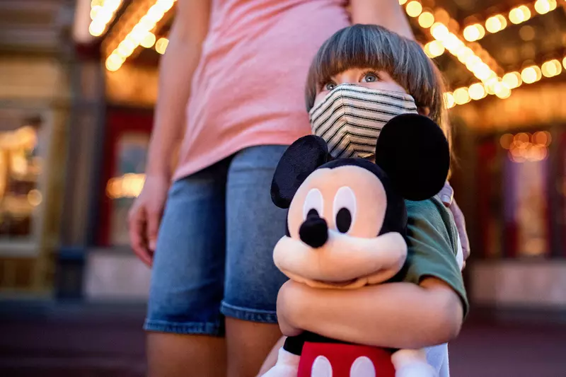 Little boy at Disney in a mask holding a stuffed Mickey Mouse.