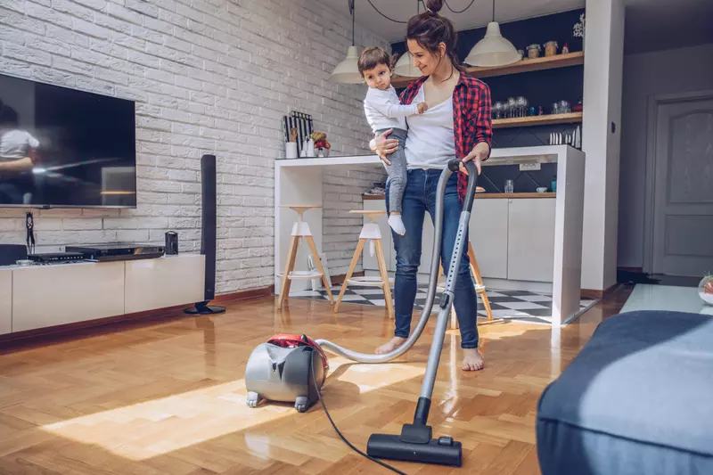 A mom juggles vacuuming the floor with holding her child at her hip.