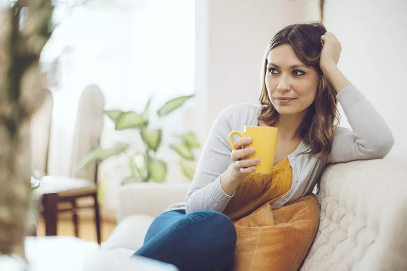 A young woman sits on her couch at home drinking her coffee