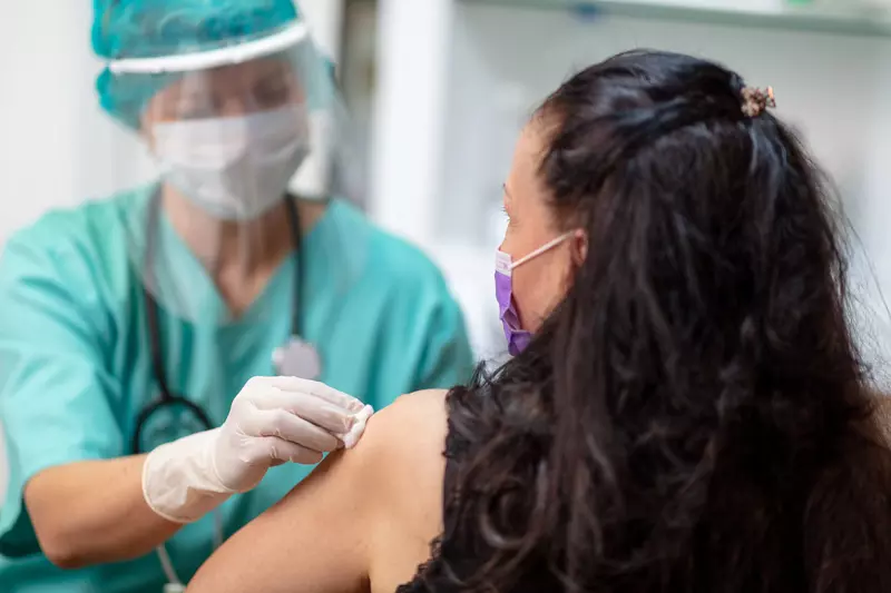 Provider prepping woman to receive a vaccine while they wear PPE and the woman wears a mask.
