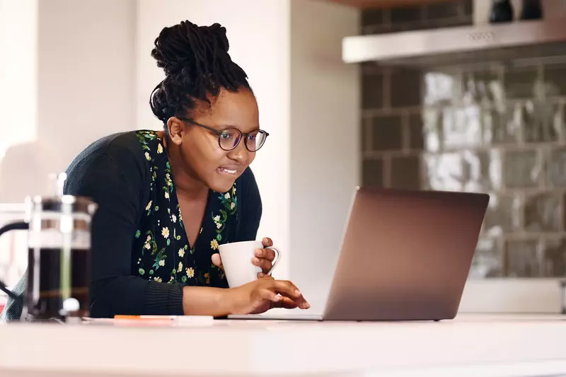 A black woman researches on her laptop while drinking coffee