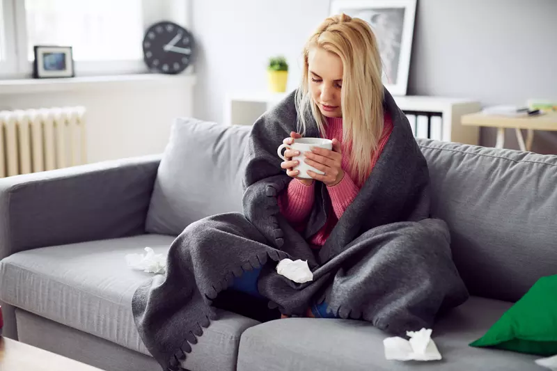 A woman drinking tea under a blanket at home. 