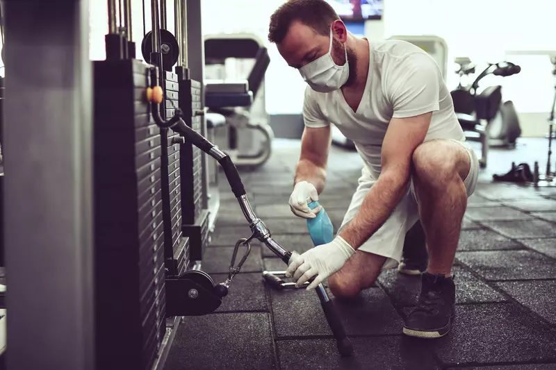 A man wearing a mask and cleaning fitness equipment in a gym.