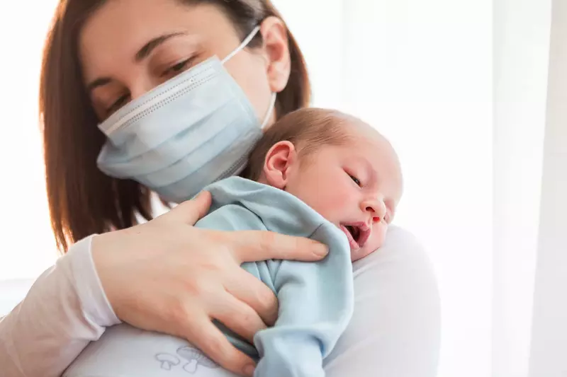 A mom wearing a mask holding her infant.