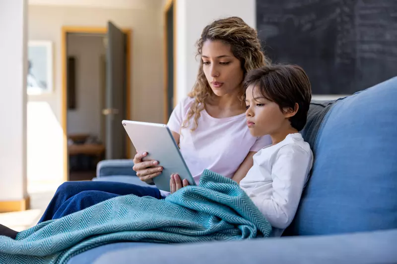 A mother and son sitting on a couch and using a tablet.