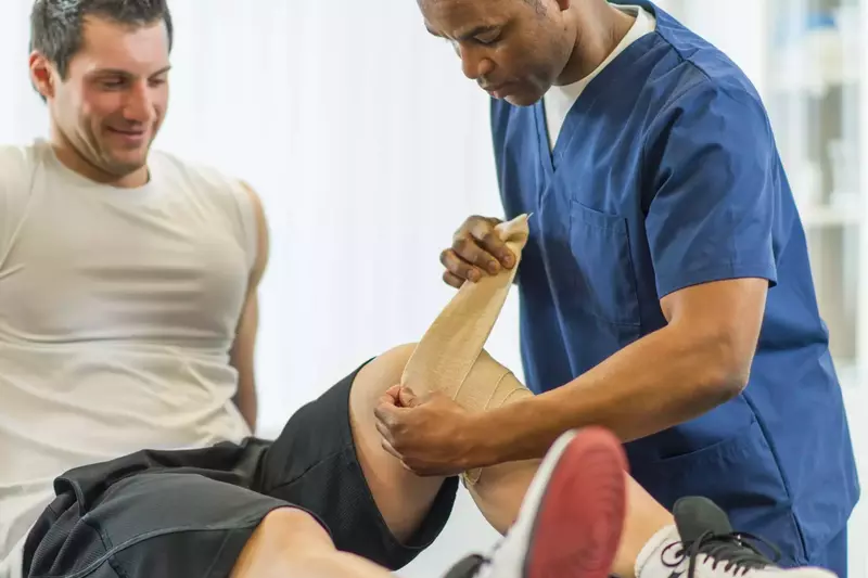 A physical therapist bandaging a male patient's left knee.