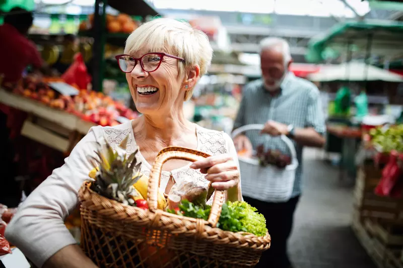Woman smiling in the foreground while her husband is in the background. Both are holding a basket of fresh produce at a farmers market.