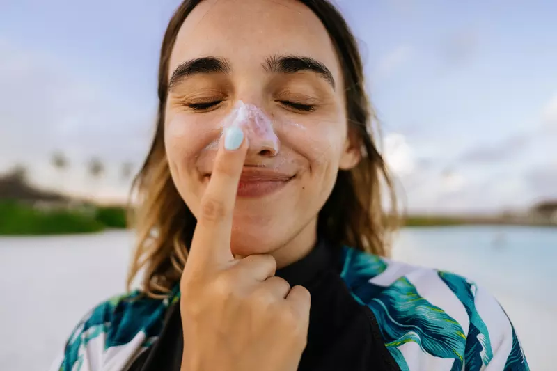 A woman on a beach wearing sunscreen on her nose and touching her nose with her index finger.