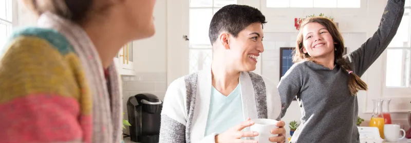 A woman dancing in the kitchen with her daughters and drinking coffee.