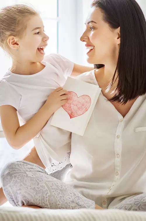 Mother and daughter sitting on a bed, holding each other with a card with a heart on it in the daughter's hands.