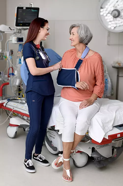 A senior woman with an arm in a sling smiling with a nurse while in an emergency room examination room.