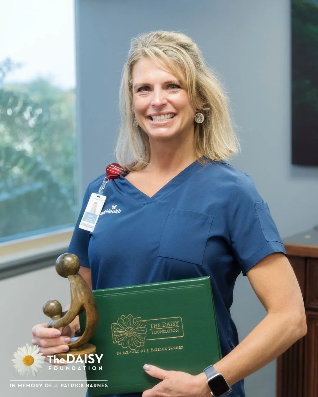 Carrie Peek, a white, blonde woman wearing blue scrubs, stands with her DAISY Award certificate and statue.