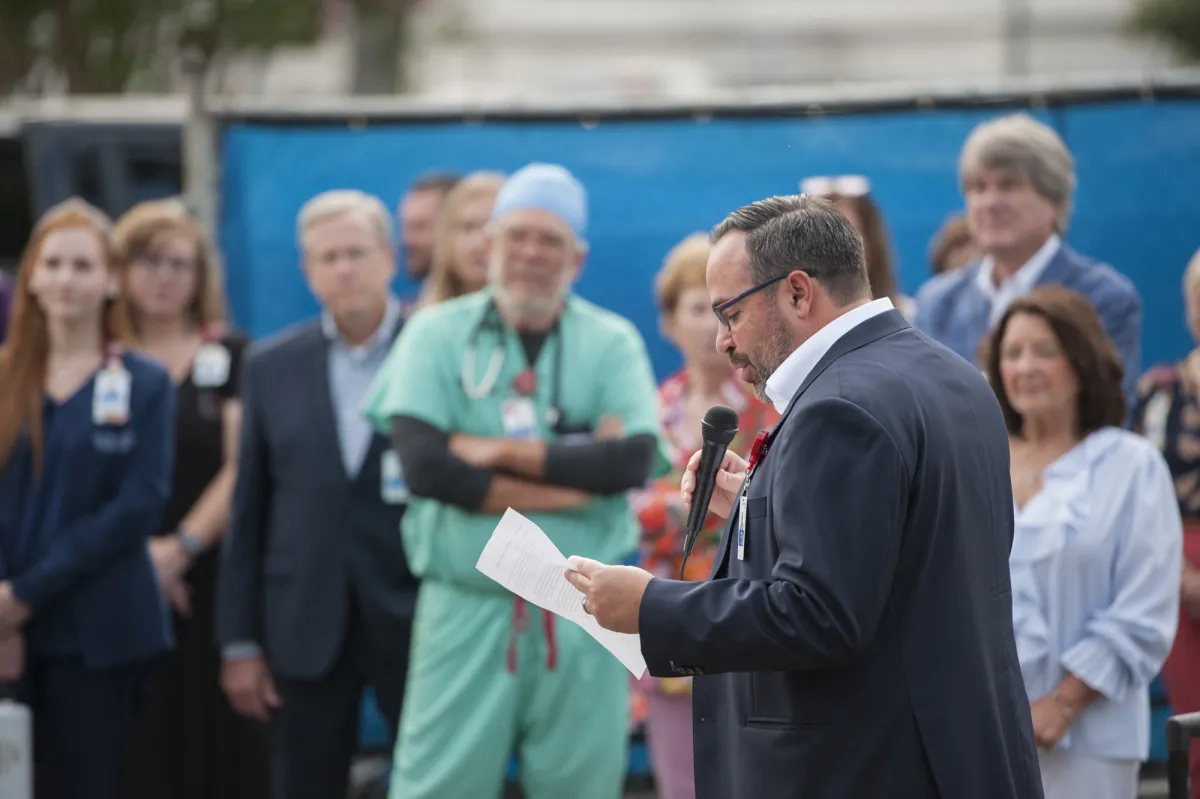 Isaac Sendros, a white man with dark hair, wearing glasses and a grey suit, speaks in front of a group of AdventHealth team members
