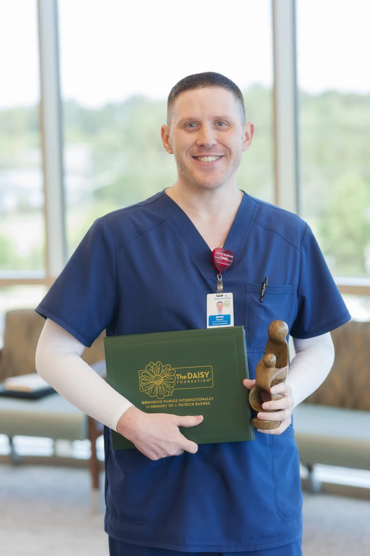 James Keeton, a white man wearing blue scrubs, stands in front of windows holding his award. 