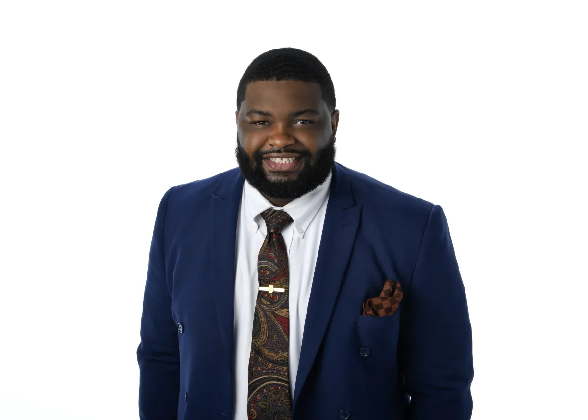 Elijah Walters, an African-American man wearing a blue suit, smiles at the camera in front of a white background