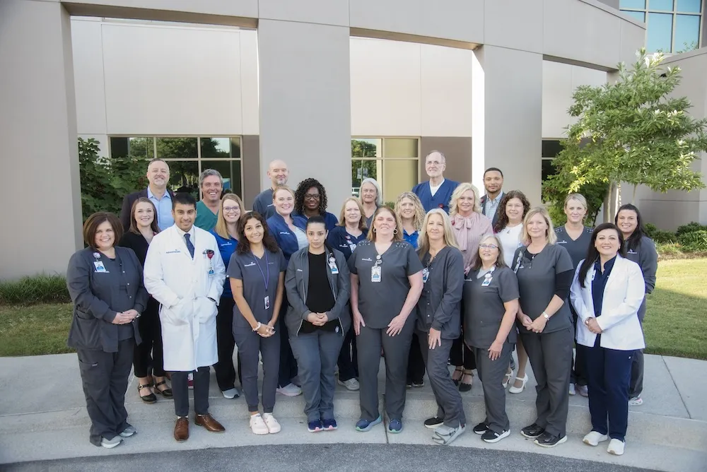 A large group of people smiling at the camera. Most are in gray scrubs, but several are wearing suits and professional attire. One individual is wearing a white doctor's coat.