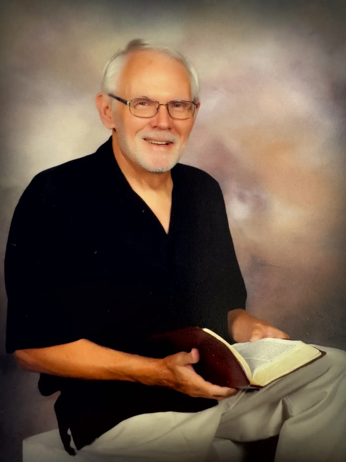 Charlie Williams sits with his Bible open in his hands in front of a beige background. He is a white man with gray hair and a gray beard. He is wearing a black shirt and khaki pants. 