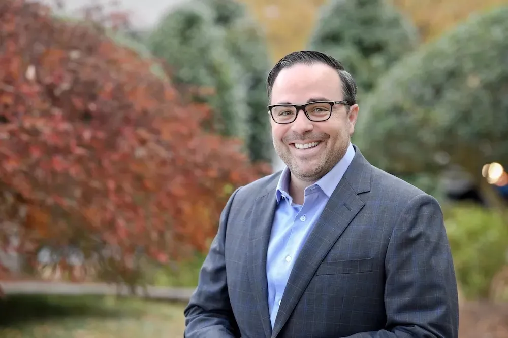 Isaac Sendros, a white man with dark hair, wearing glasses and a grey suit, stands in front of a blurred landscaped background