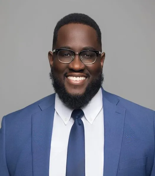 Mack Vendome, an African-American man wearing a blue suit, white shirt, and glasses, smiles in front of a plain gray background.