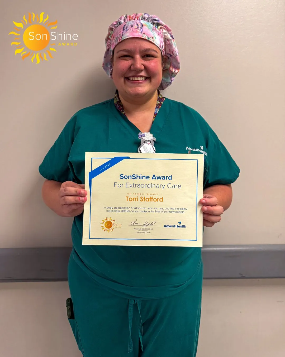 Victorria Stafford, a white woman with a pink scrub cap and teal surgical scrubs, stands in front of a beige wall holding her award certificate.