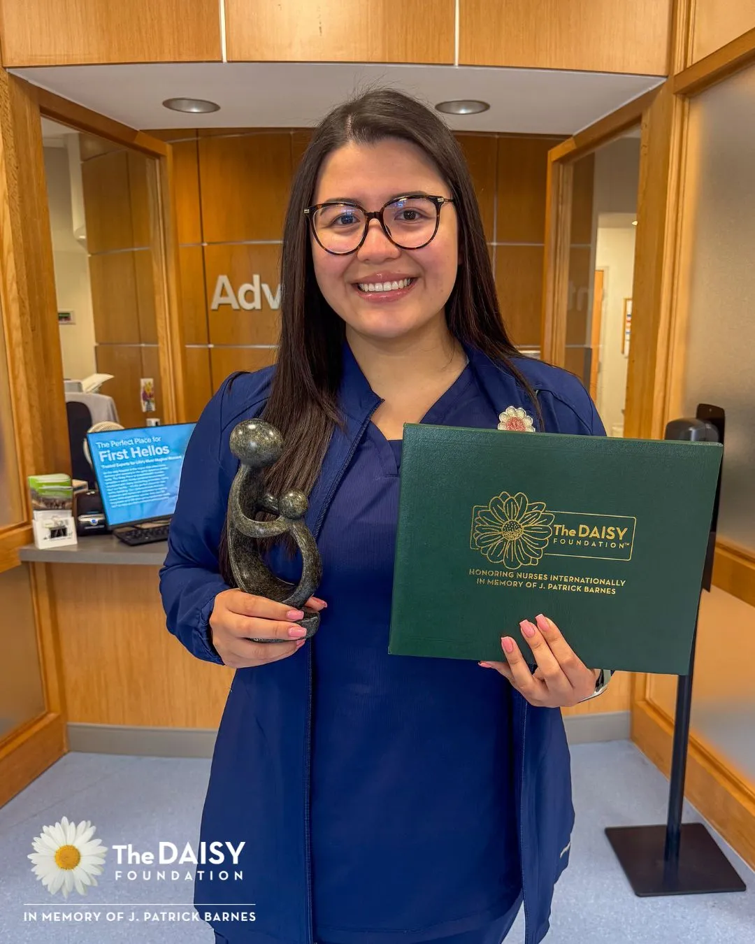 Jocelyn Villanueva, a woman with dark hair wearing navy blue scrubs, holds her DAISY Award in front of a reception area