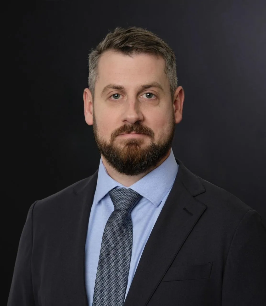 Dr. Nicholas Wells, a white, bearded brunette man, is pictured with a dark gray background. He is wearing a dark colored suit and tie with a blue collared shirt.