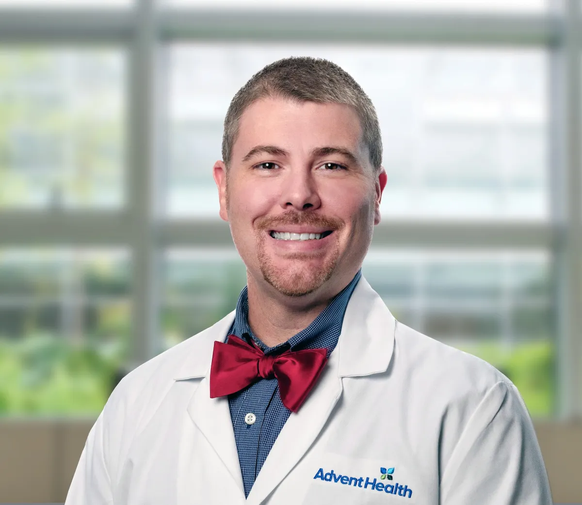 Zachary Whitener, a white man with short brown hair, stands in front of a blurred office-style background. He is wearing a blue shirt with a red bow tie and a white medical coat.