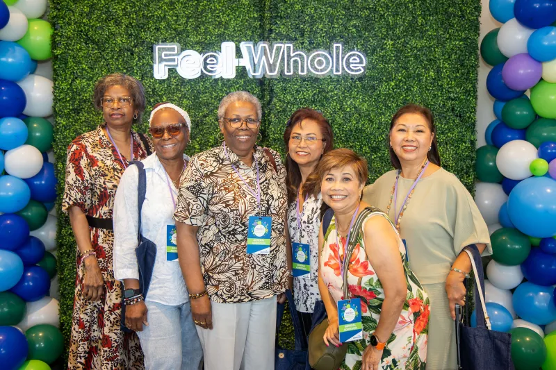 Women attending the Women’s Health and Cancer Prevention Event, taking a group picture in front of a green backdrop with a sign that states 'Feel Whole'.