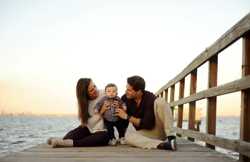 A young couple sitting on a dock holding their baby.